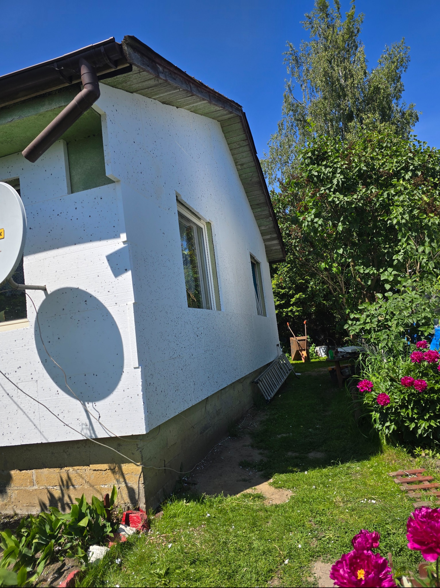 Ocieplony dom jednorodzinny z białą elewacją styropianową, widoczne okna, antena satelitarna i fragment ogrodu z kwiatami w słoneczny dzień.