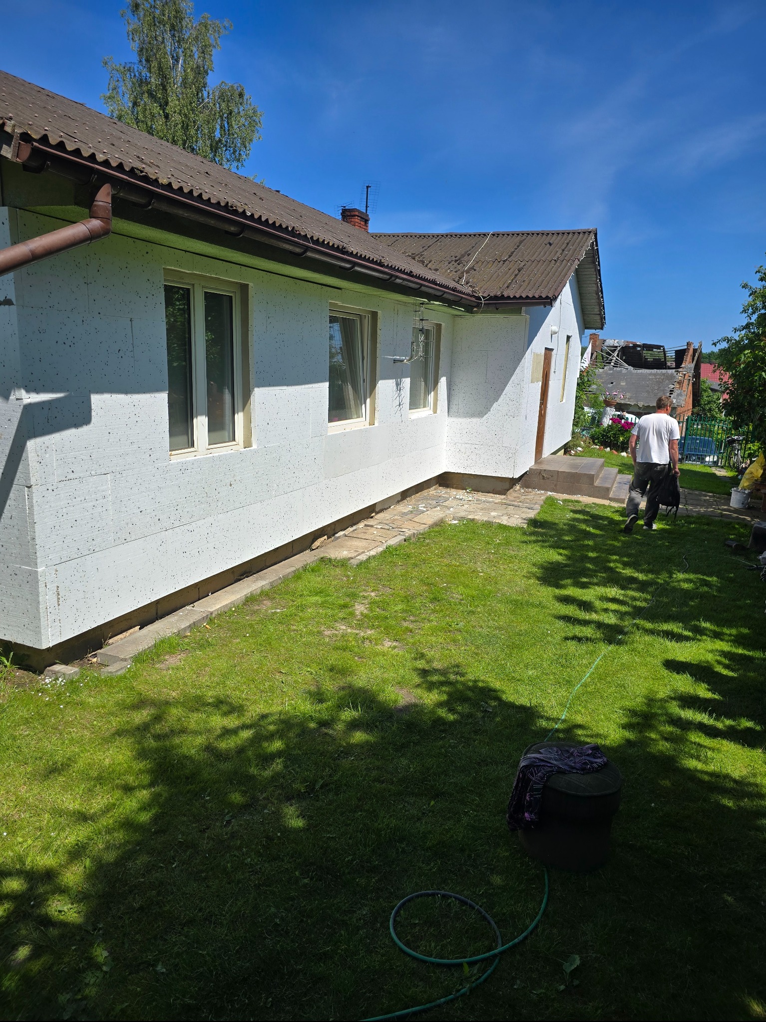 Dom z białym styropianem na elewacji, widok z zewnątrz w słoneczny dzień. Mężczyzna w tle. Prace przygotowawcze przed nałożeniem tynku.