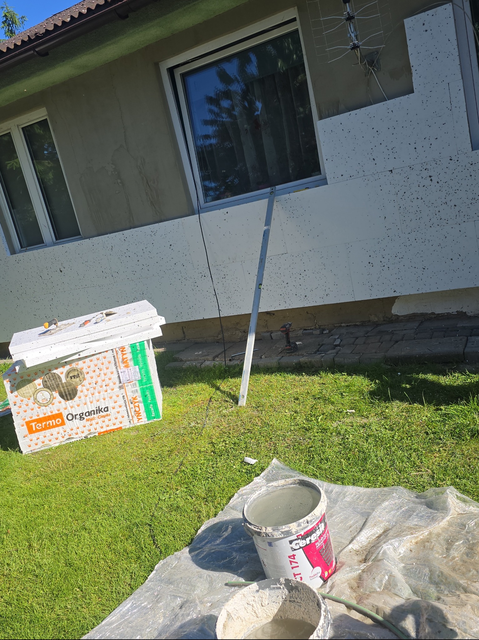 Ocieplanie elewacji styropianem: paczki styropianu Terma Organika, wiadro z klejem Ceresit CT 174 i poziomica przy ścianie budynku z ociepleniem.
