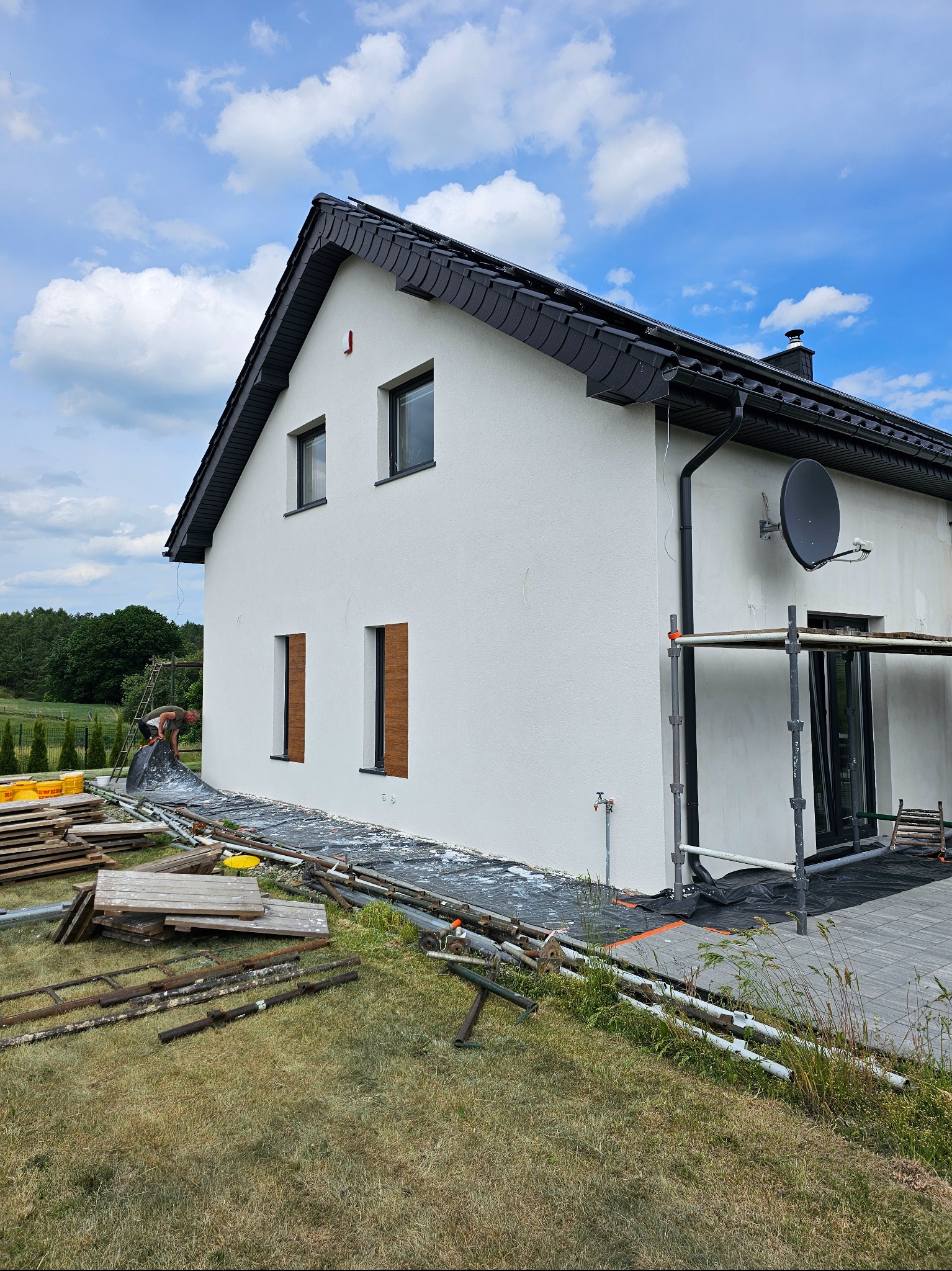Biała elewacja domu jednorodzinnego z czarnym dachem, widoczna z boku. Na trawniku leżą materiały budowlane i rusztowanie. Błękitne niebo z chmurami w tle.