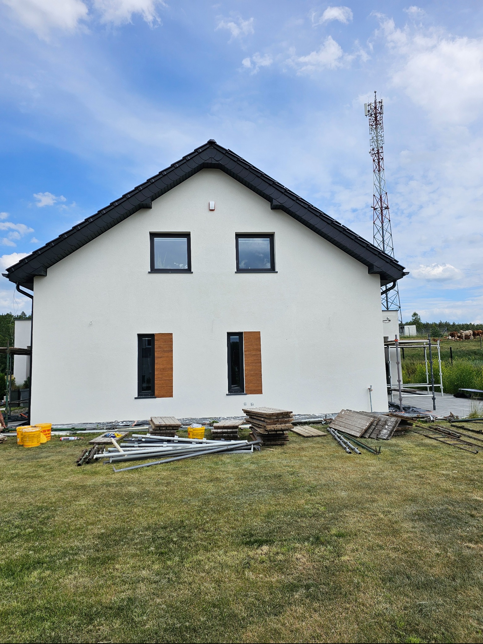 Boczny widok domu z białą elewacją i czarnym dachem. Widoczne rusztowania, materiały budowlane i wieża telekomunikacyjna w tle. Częściowo wykończona elewacja.