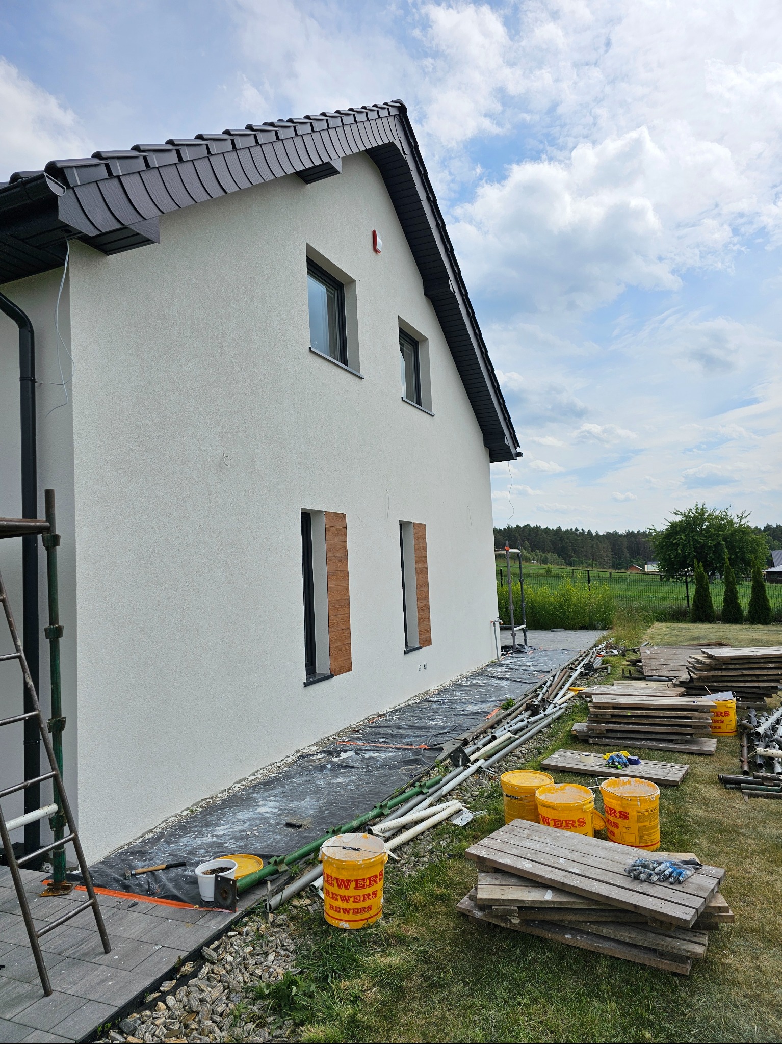 Boczny widok domu z nową, jasną elewacją. Widoczne rusztowania, narzędzia i puste otwory okienne z drewnianymi zaślepkami. Tło: zielony trawnik i błękitne niebo.