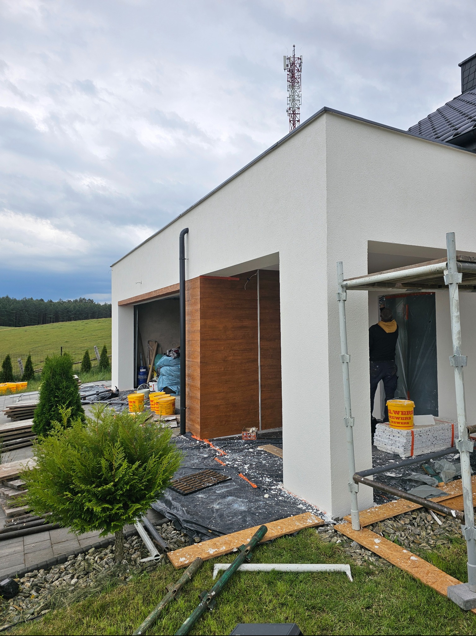 Elewacja domu jednorodzinnego w trakcie prac wykończeniowych, widoczne rusztowania, wiaderka z farbą i pracownik wewnątrz budynku. Tło: zielone wzgórza.