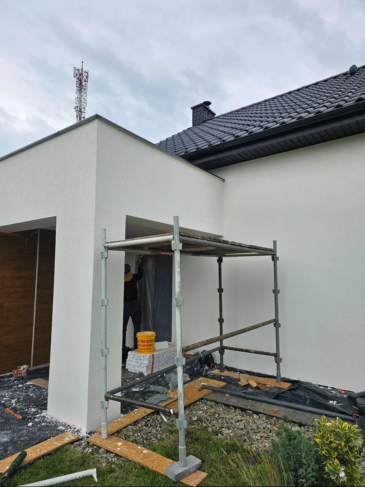 Biała elewacja domu jednorodzinnego w trakcie prac wykończeniowych, widoczne rusztowanie i pracownik w czarnym ubraniu. W tle maszt telekomunikacyjny.