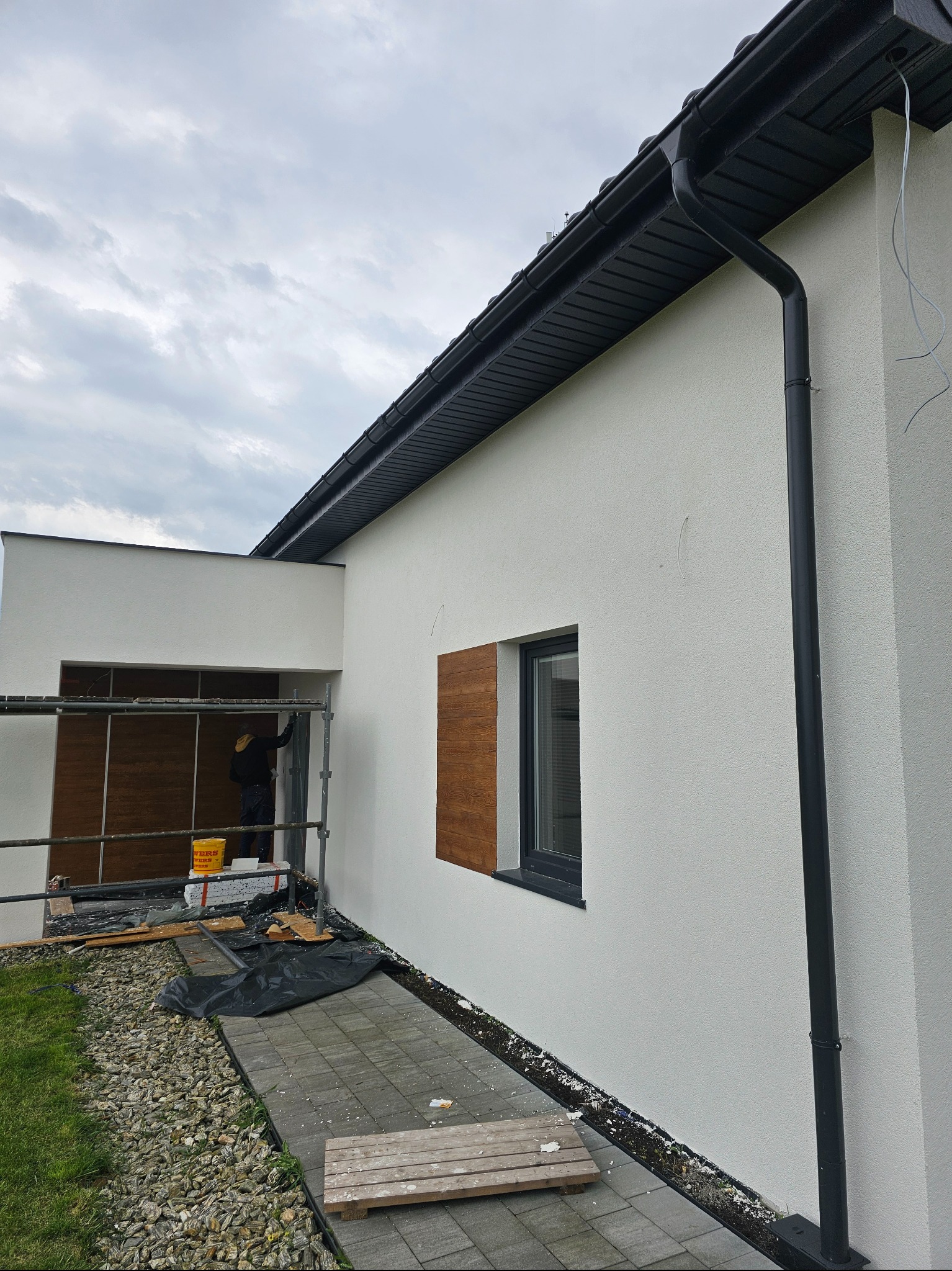 Elewacja domu jednorodzinnego w trakcie prac wykończeniowych. Widoczne rusztowanie, pracownik, fragment okna z drewnianą okiennicą i czarna rynna.