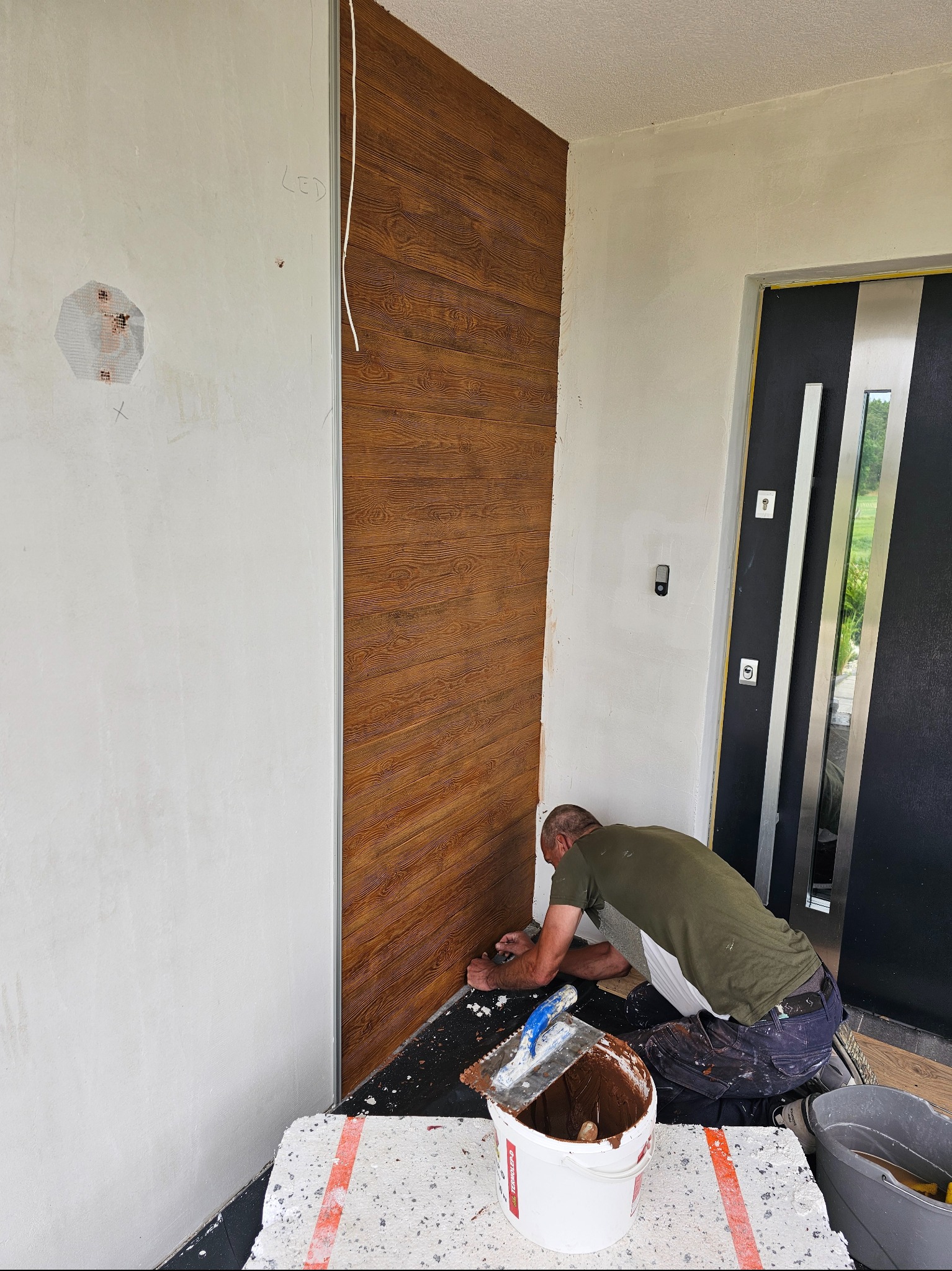 Pracownik klei panel dekoracyjny imitujący drewno na ścianie przy wejściu do domu. Widoczne narzędzia i wiadro z klejem. Fragment drzwi wejściowych.
