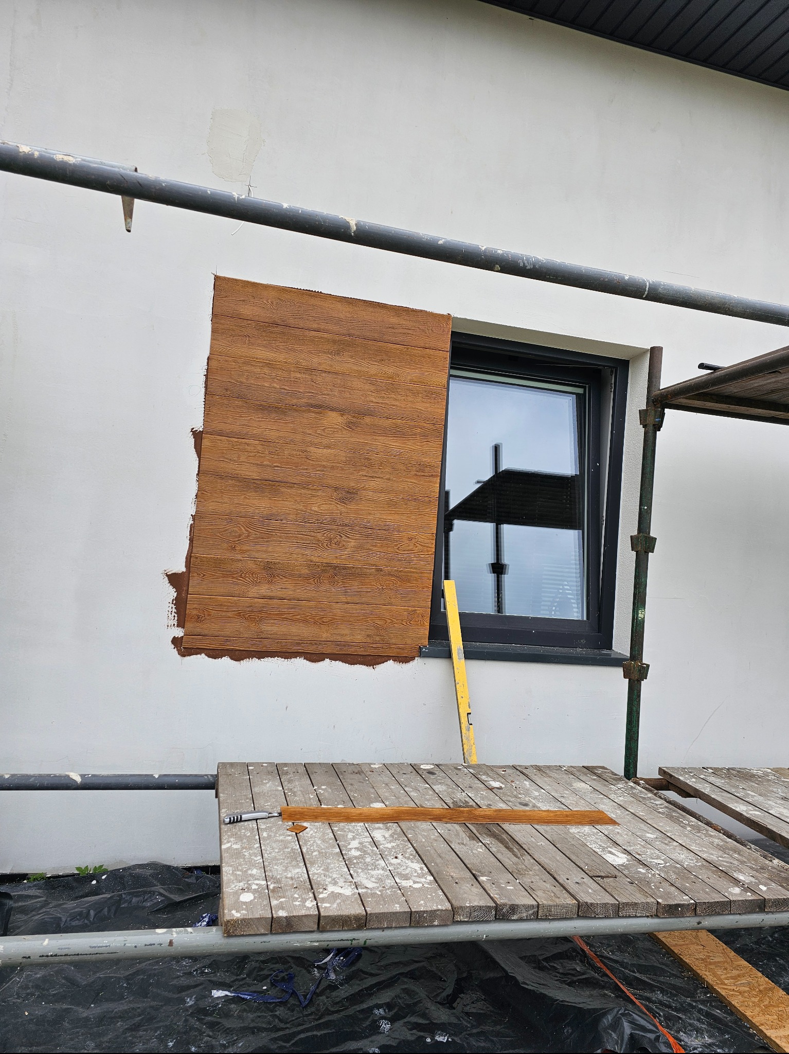 Montaż paneli elewacyjnych imitujących drewno na białej ścianie budynku, widoczne rusztowanie i narzędzia, etap prac przy oknie.