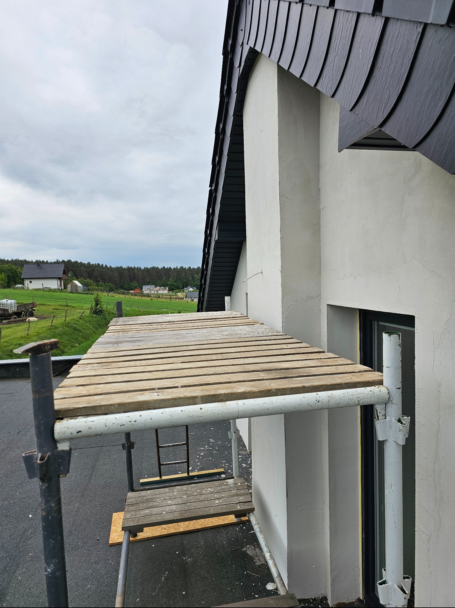 Elewacja domu w trakcie prac wykończeniowych, widok z rusztowania. Świeżo otynkowana ściana, fragment dachu pokrytego łupkiem. W tle zielony krajobraz.