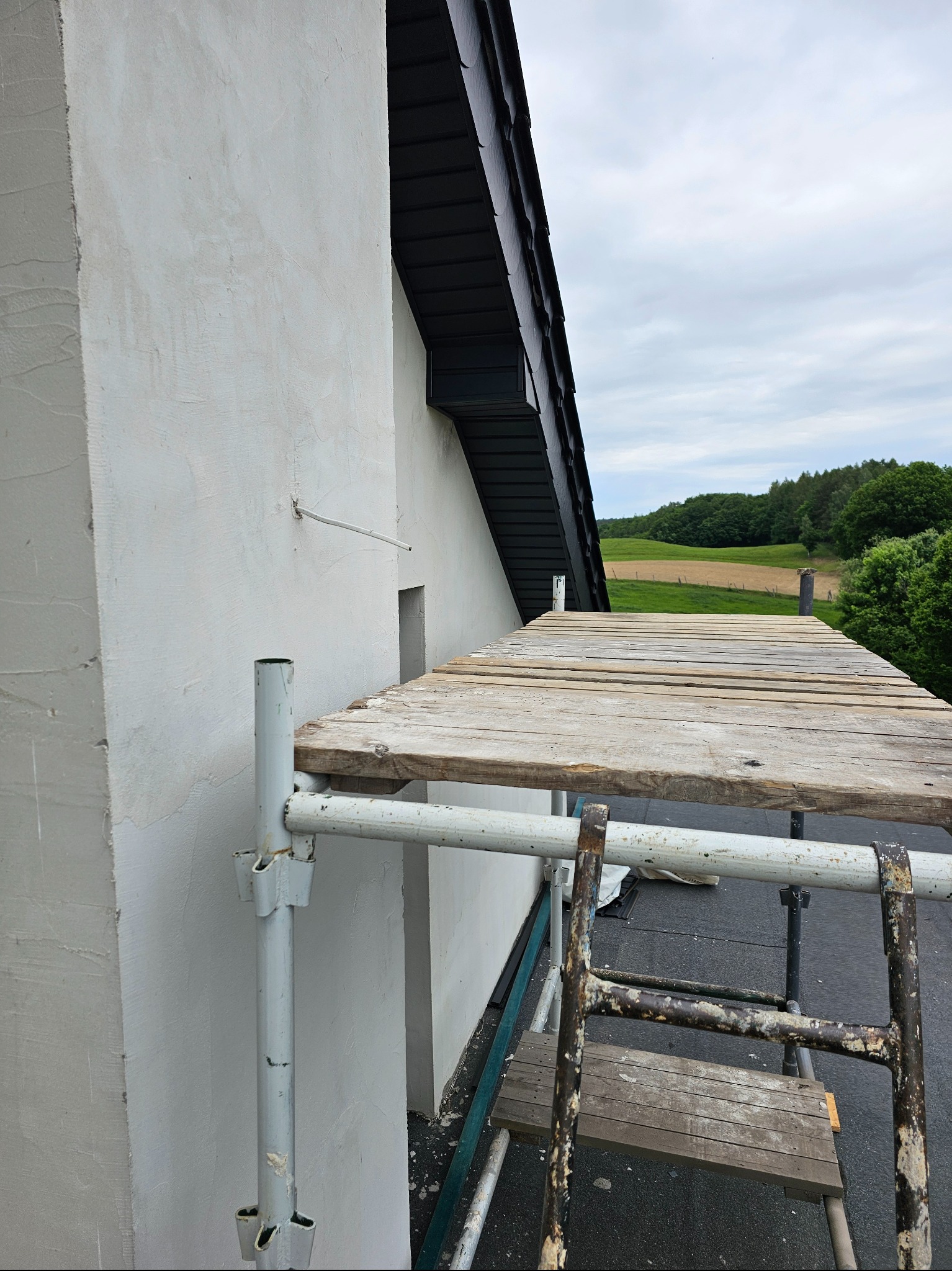 Ściana budynku po szpachlowaniu, widoczne rusztowanie i drabina. Tło stanowią pola i lasy pod pochmurnym niebem.