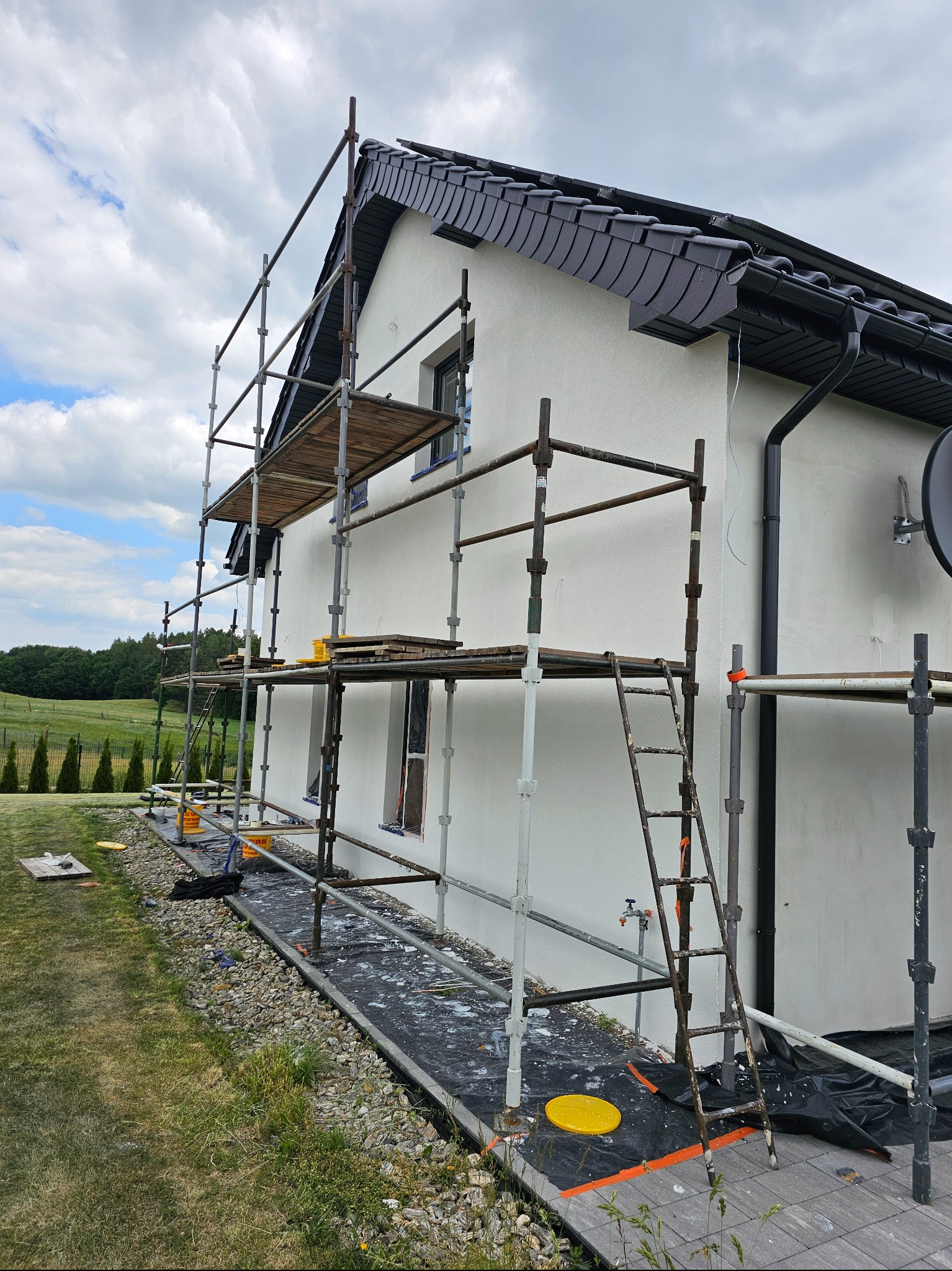 Biały dom jednorodzinny w trakcie prac elewacyjnych, otoczony rusztowaniem. Widoczne narzędzia i materiały budowlane. Tło stanowi zielony krajobraz.