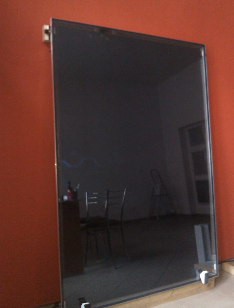 Czarny panel słoneczny w trakcie montażu, oparty o pomarańczową ścianę, z widocznym odbiciem wnętrza pokoju i mebli.