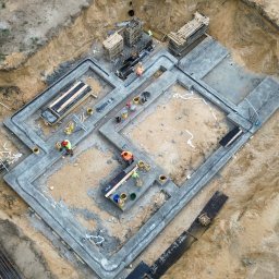Budowa po inżyniersku - Z góry widok na wylane fundamenty domu jednorodzinnego. Widoczni pracownicy w odblaskowych kamizelkach i kaskach. Na ziemi leżą narzędzia i materiały budowlane.