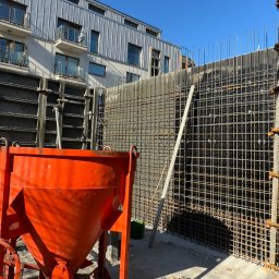 Budowa po inżyniersku - Pomarańczowy pojemnik na beton na tle zbrojenia ściany i szalunków. W tle budynek mieszkalny z balkonami i metalowym dachem. Słoneczny dzień.
