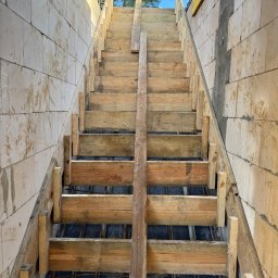 Budowa po inżyniersku - Szczegółowe ujęcie szalunku schodów z drewnianych desek, zbrojenia i bloczków betonowych, w trakcie budowy. Widok z dołu, niebo w tle.