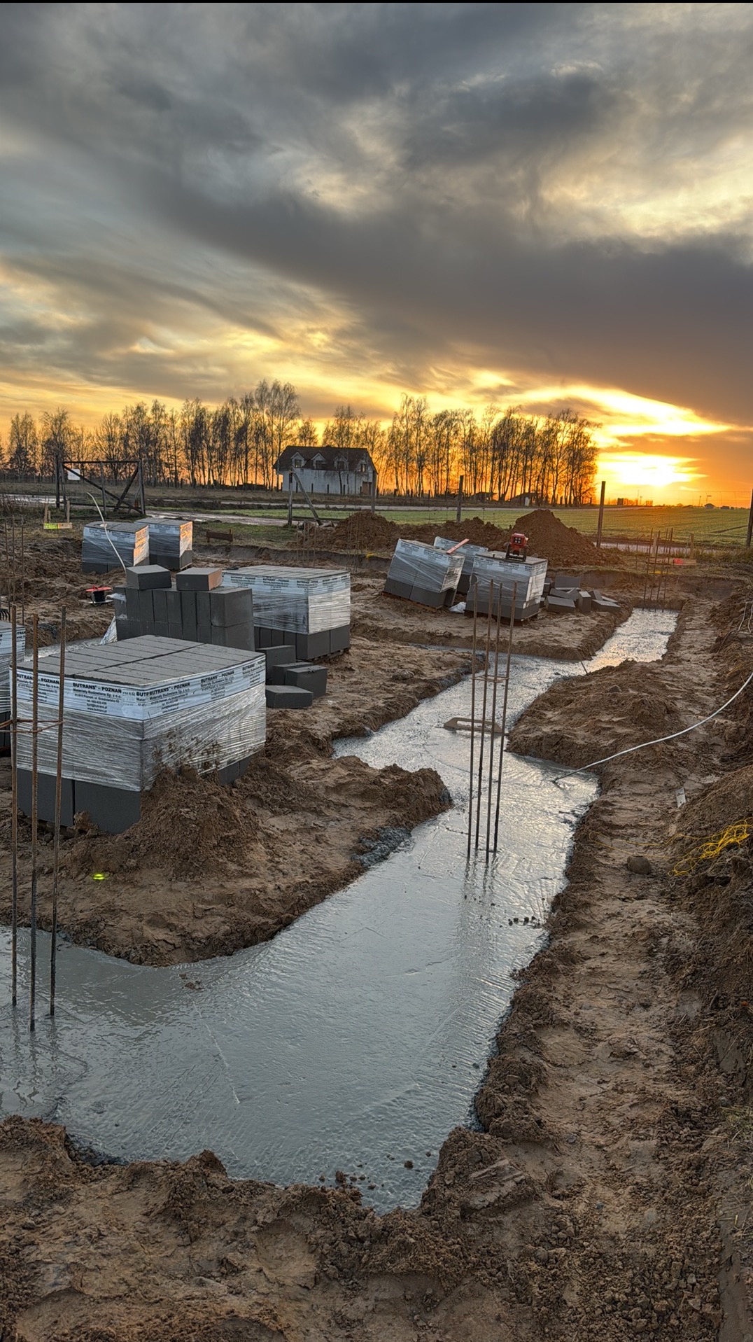 Zalane fundamenty pod budowę domu o zachodzie słońca. Widoczne zbrojenia, bloczki fundamentowe w folii i budynek w tle. Ujęcie z poziomu gruntu.