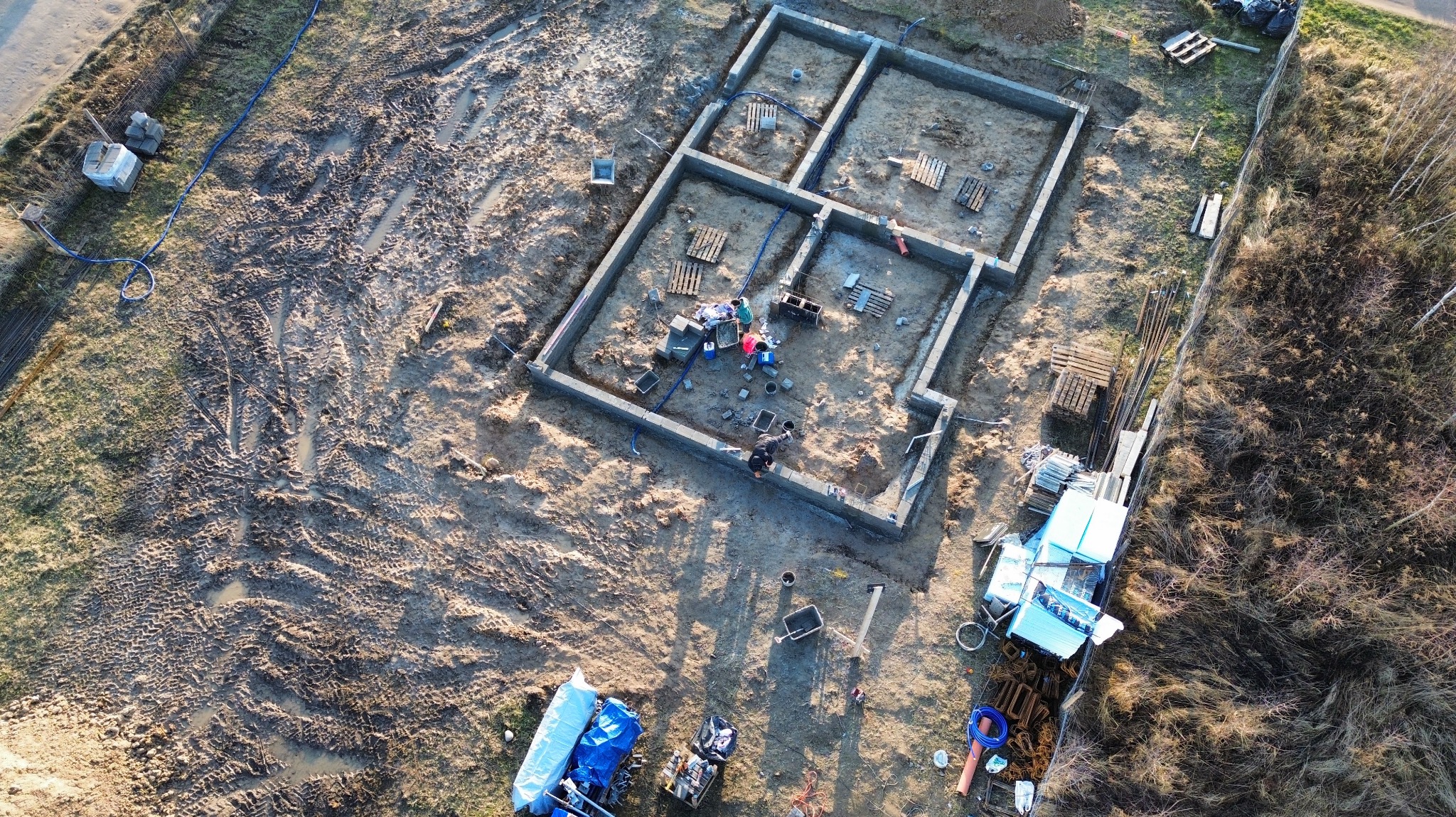 Z lotu ptaka: wylane fundamenty domu z podziałem na pomieszczenia, widoczne palety, narzędzia i ekipa budowlana przy pracy. Teren wokół rozjeżdżony.