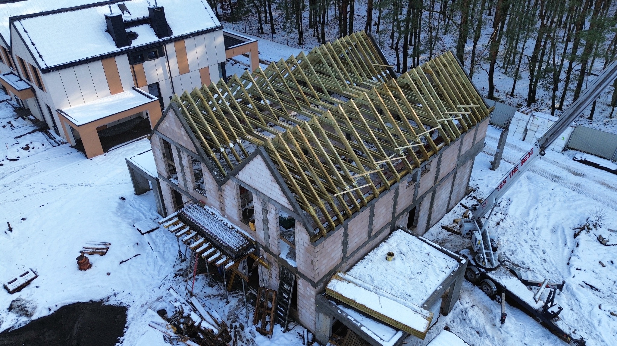 Budowa domu w zimowej scenerii: surowe mury z keramzytu, drewniana konstrukcja dachu i dźwig na ośnieżonym terenie. Widok z góry ukazuje postęp prac.