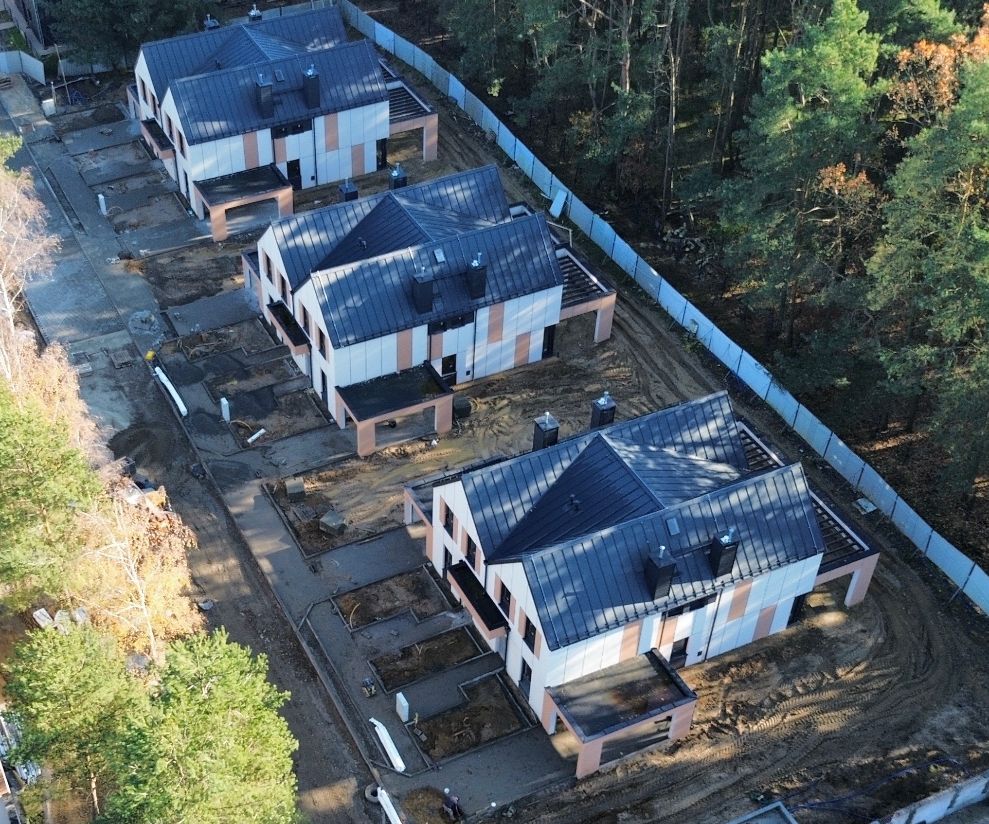 Z lotu ptaka: trzy nowoczesne domy w zabudowie szeregowej, z ciemnymi dachami i jasnymi elewacjami z akcentami w kolorze terakoty, otoczone zielenią i ogrodzeniem.