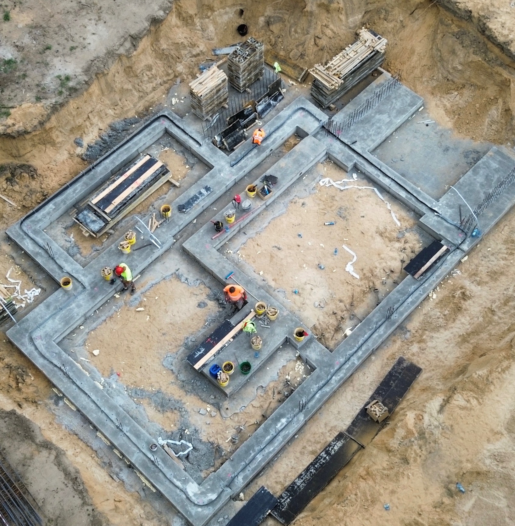 Z góry widok na wylane fundamenty domu jednorodzinnego. Widoczni pracownicy w odblaskowych kamizelkach i kaskach. Na ziemi leżą narzędzia i materiały budowlane.