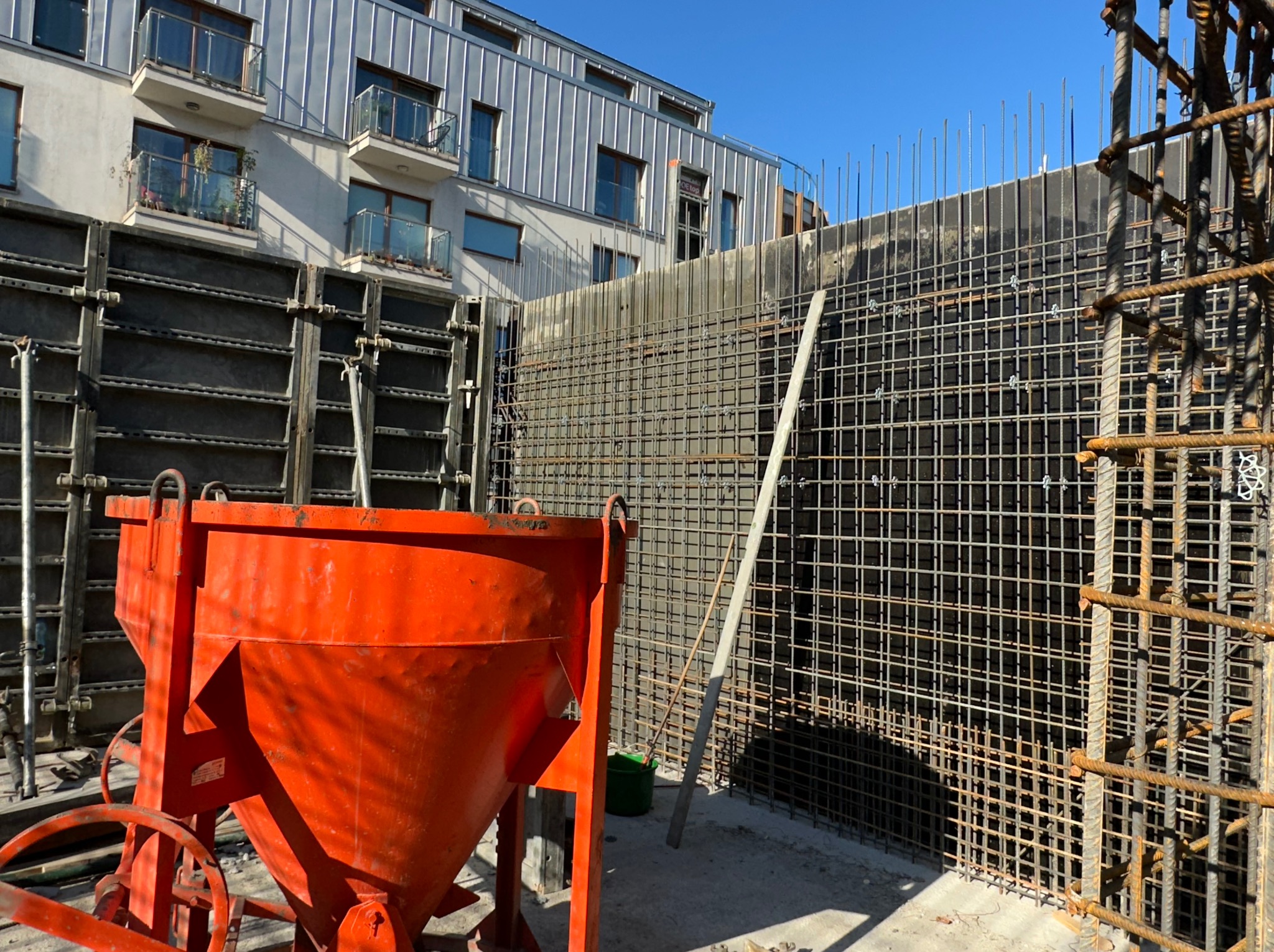 Pomarańczowy pojemnik na beton na tle zbrojenia ściany i szalunków. W tle budynek mieszkalny z balkonami i metalowym dachem. Słoneczny dzień.