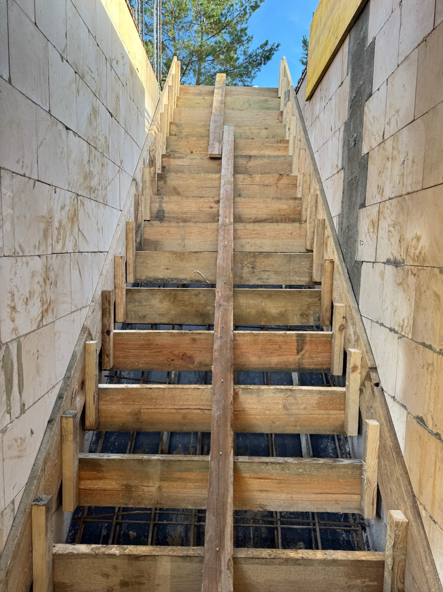 Szczegółowe ujęcie szalunku schodów z drewnianych desek, zbrojenia i bloczków betonowych, w trakcie budowy. Widok z dołu, niebo w tle.