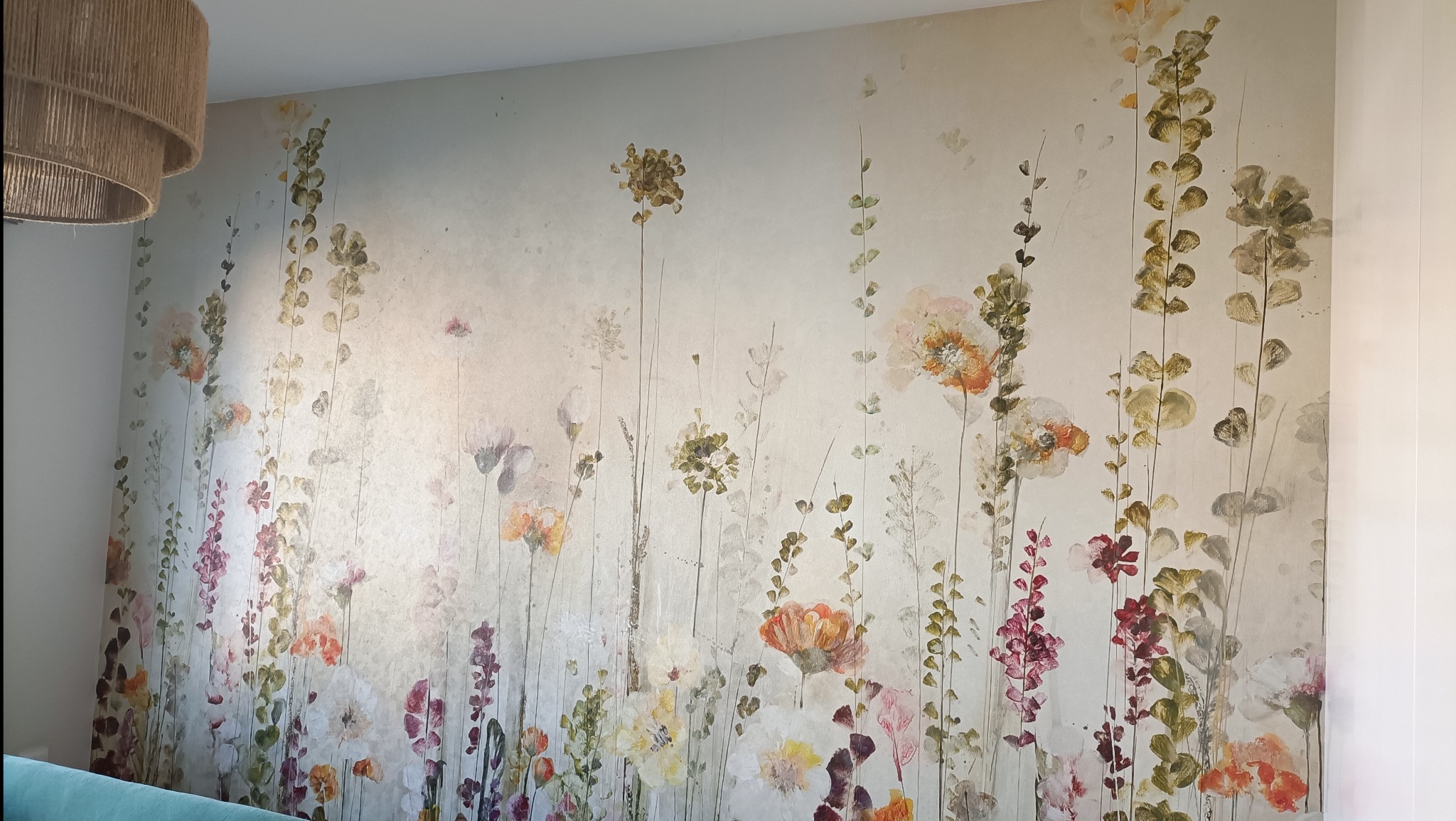 Ściana z dekoracyjną tapetą w kwiaty w pastelowych barwach, widoczny fragment lampy z naturalnego sznurka. Subtelne wykończenie wnętrza.
