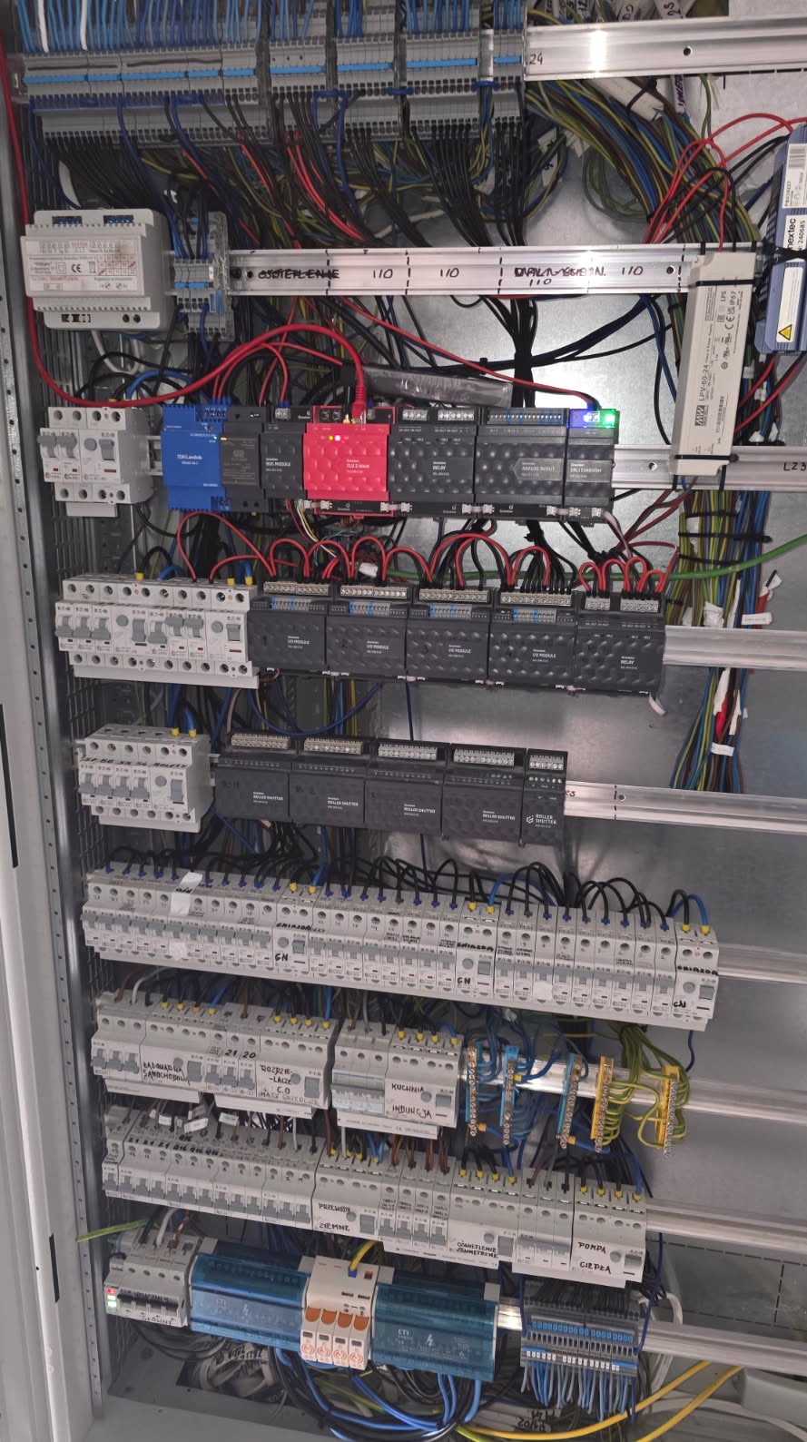 Rozbudowana szafa sterownicza z modułami, przewodami i zabezpieczeniami, widoczne oznaczenia stref,  różne kolory modułów (czerwony, niebieski, czarny).