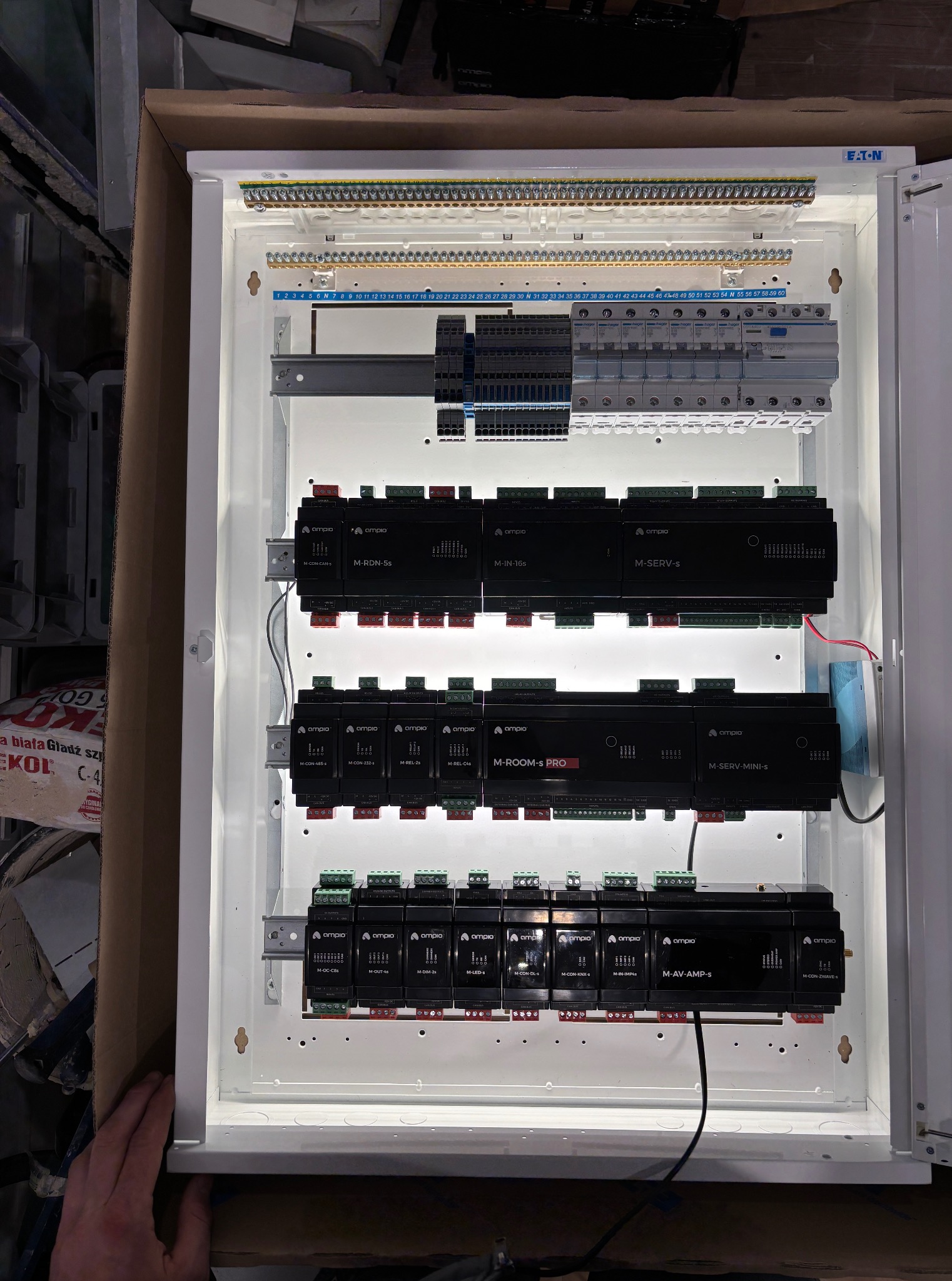 Rozdzielnica elektryczna z modułami sterującymi Ampio, widoczne liczne złącza i przewody. Całość oświetlona, w białej obudowie, z elementami automatyki domowej.