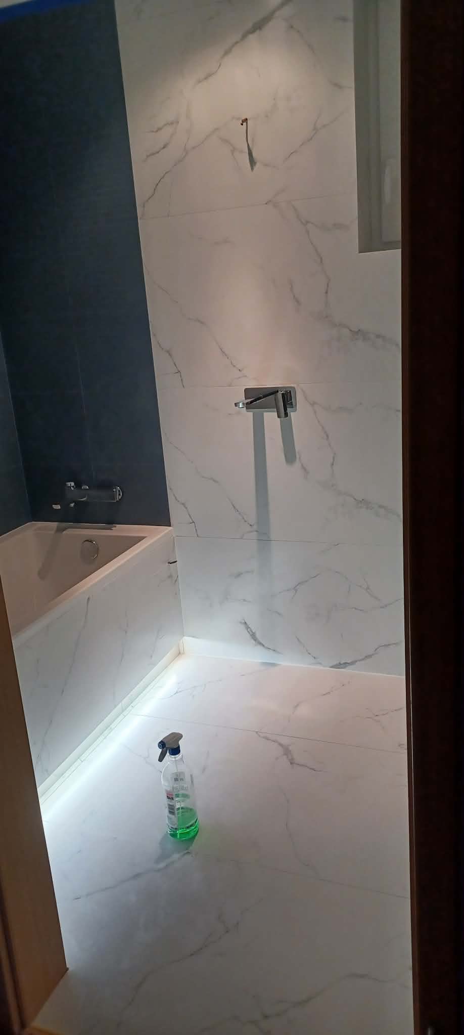 Wykończona łazienka z białymi płytkami imitującymi marmur i ciemnoniebieską ścianą. Widoczna wanna z podświetleniem LED i butelka środka czyszczącego na podłodze.