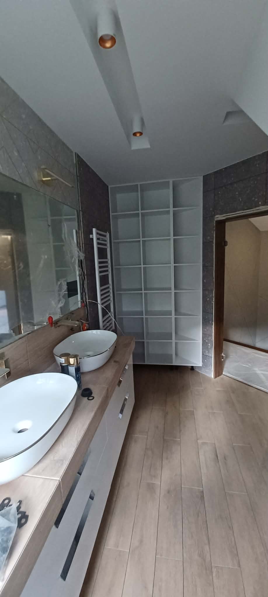 Wykończona łazienka z dwiema umywalkami nablatowymi, grzejnikiem drabinkowym i regałem w zabudowie. Na podłodze panele imitujące drewno.