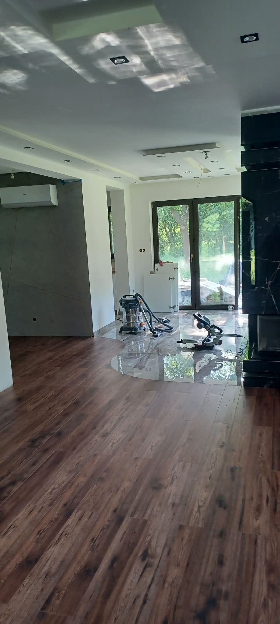 Wnętrze w trakcie remontu: parkiet ułożony, widoczny odkurzacz przemysłowy i piła do cięcia paneli. Świeżo pomalowane ściany i sufit z oświetleniem punktowym.