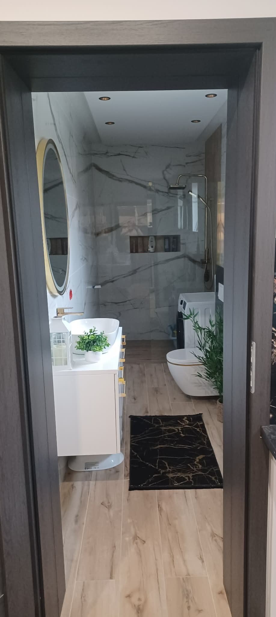 Nowoczesna łazienka z marmurowymi płytkami, kabiną prysznicową, umywalką z szafką i okrągłym lustrem w złotej ramie. Widok zza futryny drzwi.