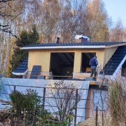 VERTIKAL PLUS OLEH KOLOBOV - Pracownik na drabinie montuje dachówkę na dobudówce domu z oknami bez szyb. Częściowo ukończony dach z czarną dachówką. W tle drzewa i niebieskie niebo.