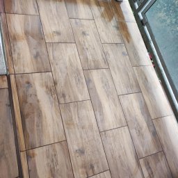 VERTIKAL PLUS OLEH KOLOBOV - Ułożona, brązowa terakota imitująca drewno na balkonie z metalową barierką i widokiem na zewnątrz. Płytki ułożone w prostokątnym wzorze, lekko połyskujące.