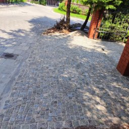VERTIKAL PLUS OLEH KOLOBOV - Ułożona kostka granitowa tworząca ścieżkę w ogrodzie, widoczna brama z ceglanymi słupkami i drzewa. Kostka w odcieniach szarości, oświetlona słońcem, z widocznymi cieniami drzew.