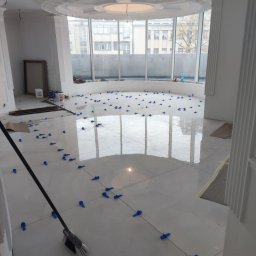 VERTIKAL PLUS OLEH KOLOBOV - Błyszcząca, jasna podłoga z płytek w trakcie układania, z niebieskimi krzyżykami dystansowymi. Widok na okno i fragment sufitu z dekoracyjnym elementem.