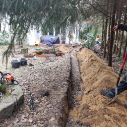 VERTIKAL PLUS OLEH KOLOBOV - Wykopany rów z drenażem wypełniony żwirem, widoczna ziemia, drzewa i narzędzia. Prace ziemne w ogrodzie, przygotowanie do odprowadzania wody.