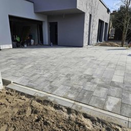 VERTIKAL PLUS OLEH KOLOBOV - Ułożona szara kostka brukowa na podjeździe nowoczesnego domu. Widoczny fragment elewacji, skrzynka elektryczna i pracownik zamiatający powierzchnię.