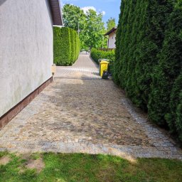 VERTIKAL PLUS OLEH KOLOBOV - Ułożona kostka granitowa tworząca ścieżkę między budynkiem a żywopłotem z tui, z żółtym pojemnikiem na śmieci w tle. Widok z poziomu trawnika.