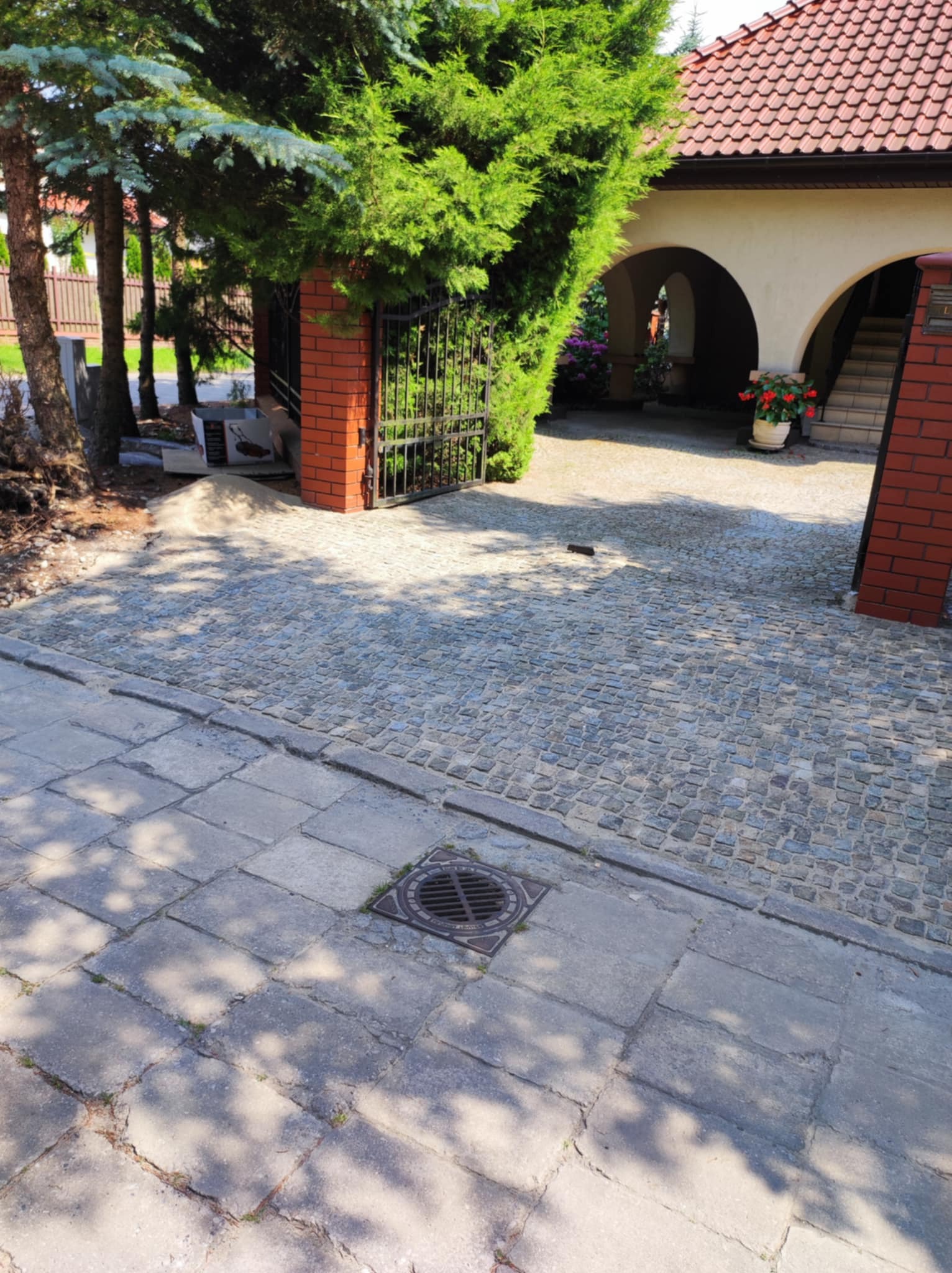 Ułożona kostka granitowa na podjeździe, widok na bramę z ceglanymi słupkami i zadaszone wejście do domu. W tle zieleń drzew i krzewów.