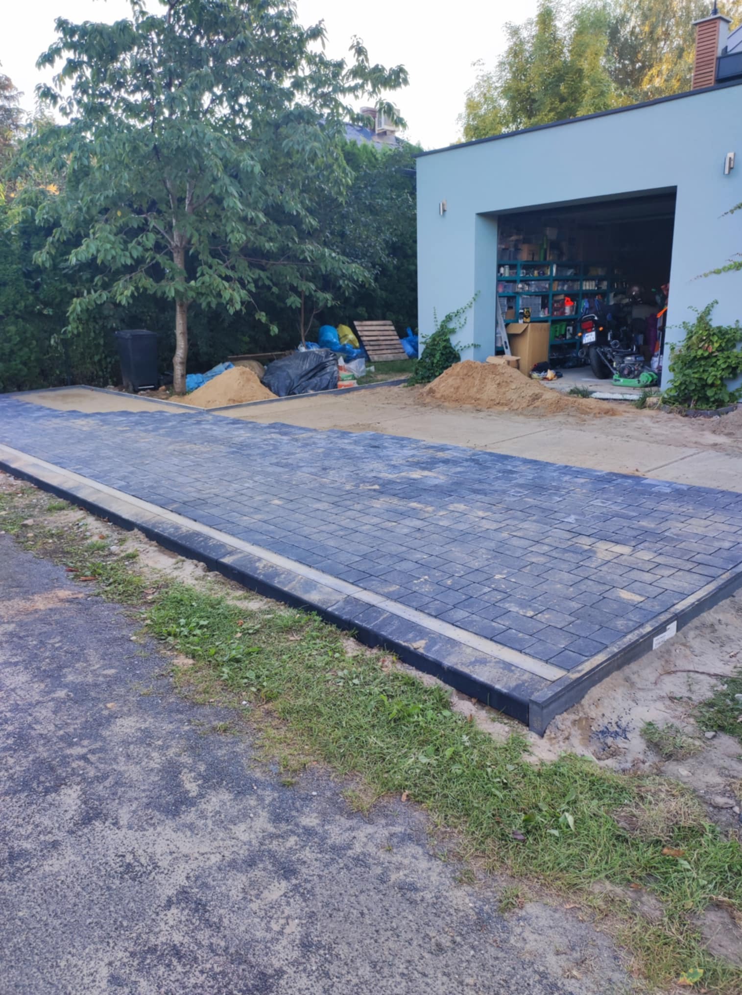 Ułożona, szara kostka brukowa z czarnym obrzeżem na podjeździe, z widocznym garażem w tle i przygotowanym piaskiem do zasypania szczelin.