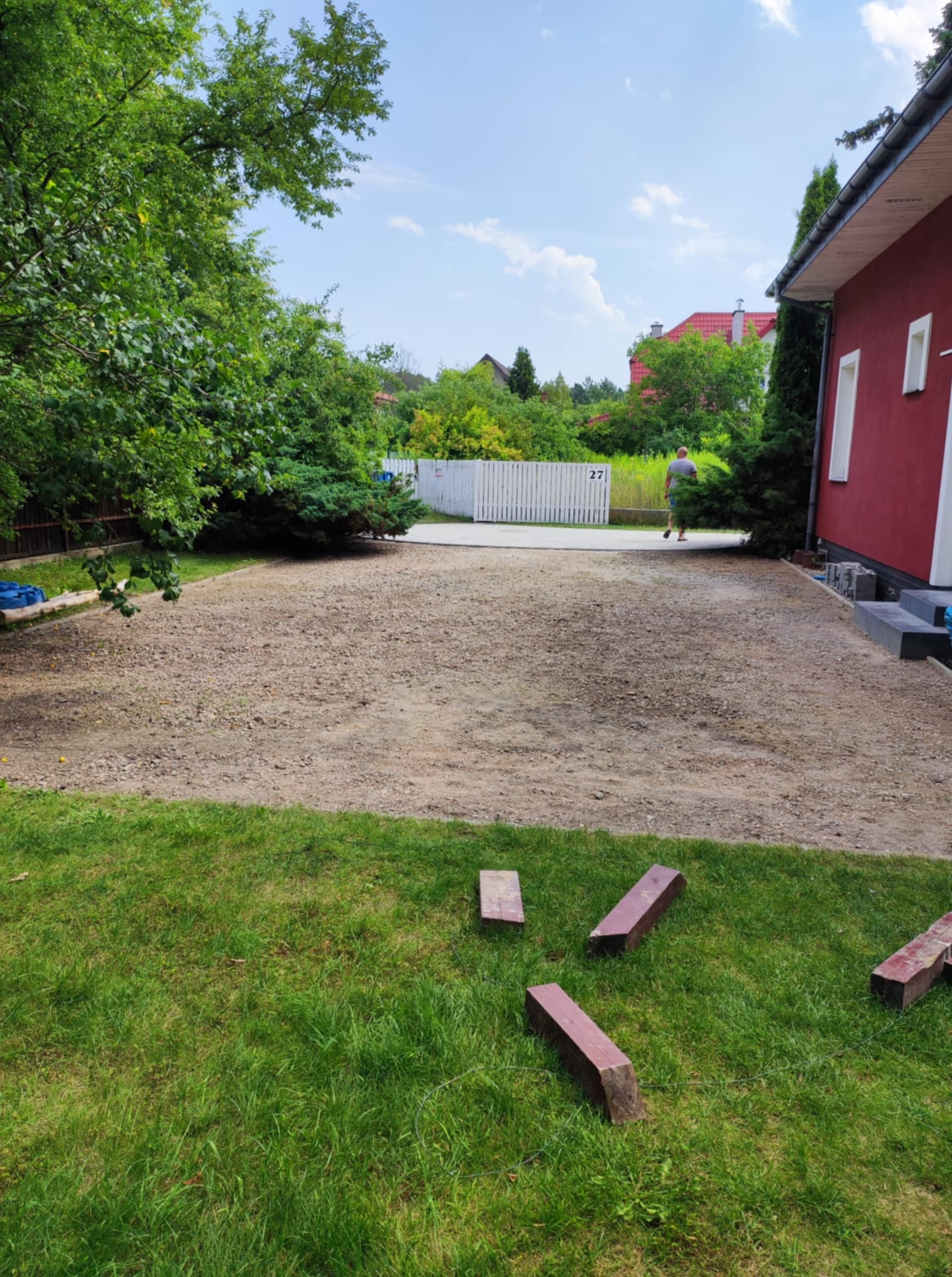 Przygotowany teren pod budowę z widocznymi cegłami na trawie, obok czerwony budynek i białe ogrodzenie w tle, w słoneczny dzień.