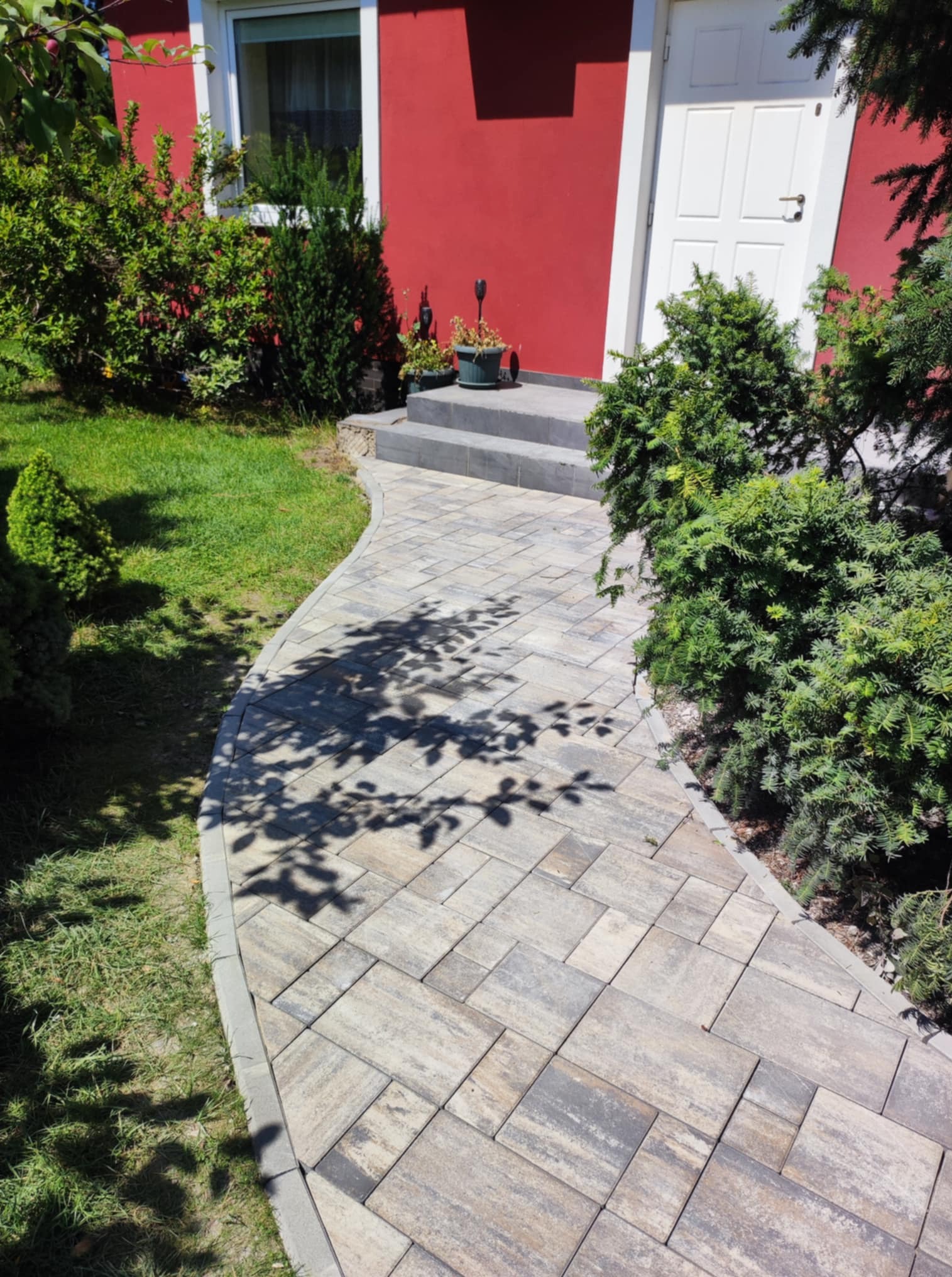 Zakrzywiony chodnik z kostki brukowej prowadzi do domu z czerwonymi ścianami i białymi drzwiami, otoczony zielenią. Cień drzewa pada na ścieżkę.
