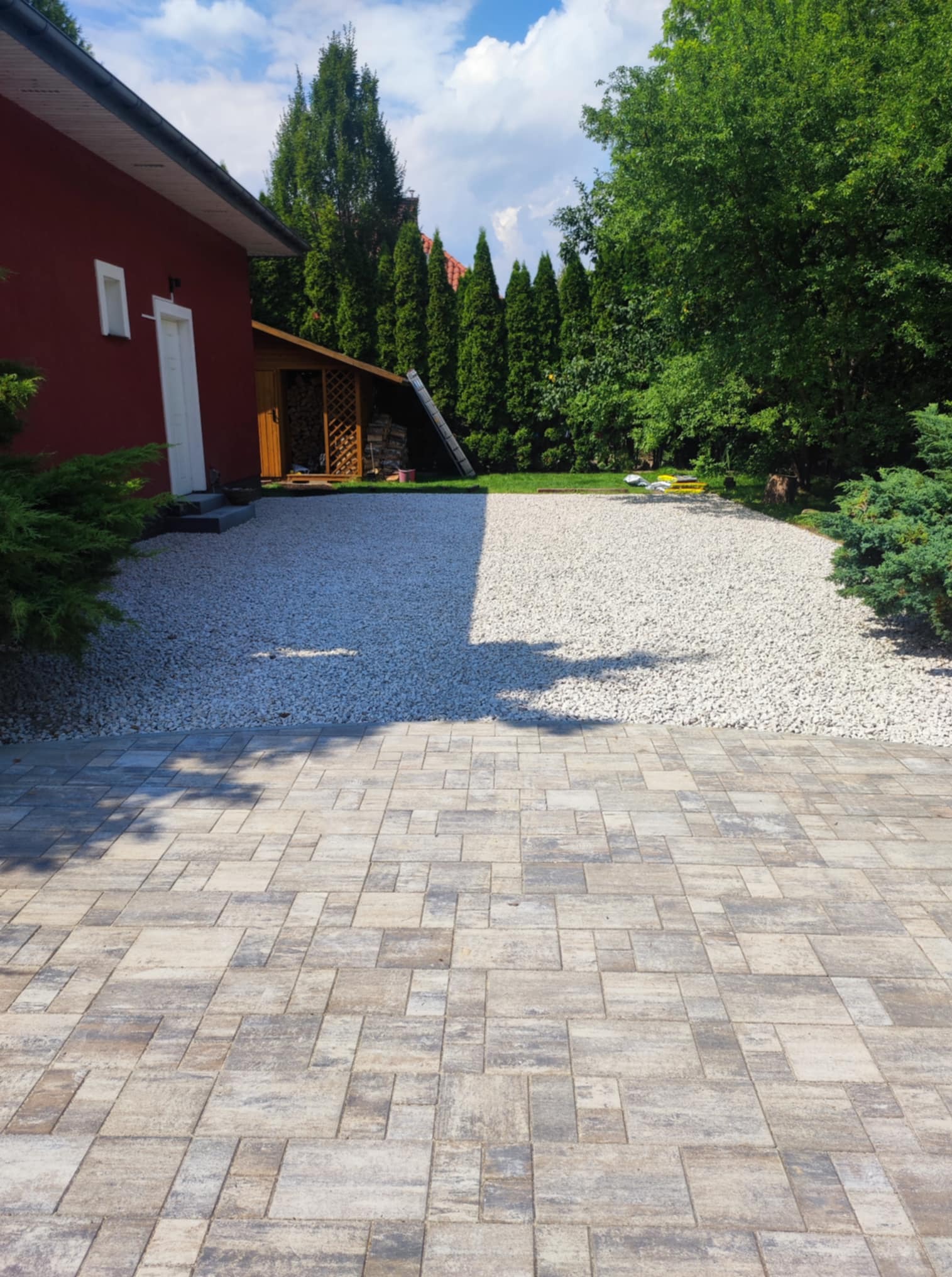 Ułożona kostka brukowa w odcieniach szarości i beżu, przechodząca w jasny żwir. W tle budynek z czerwoną elewacją i zielone drzewa.