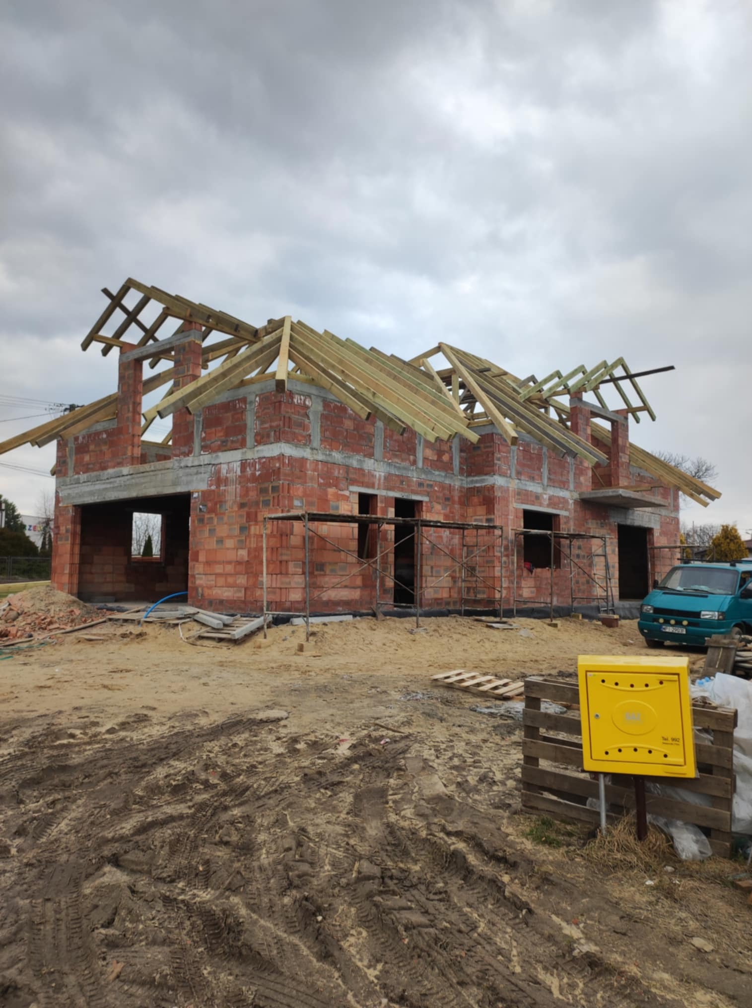 Surowy stan domu z czerwonej cegły z drewnianą konstrukcją dachu w trakcie budowy, na zewnątrz widoczny teren budowy i skrzynka gazowa.
