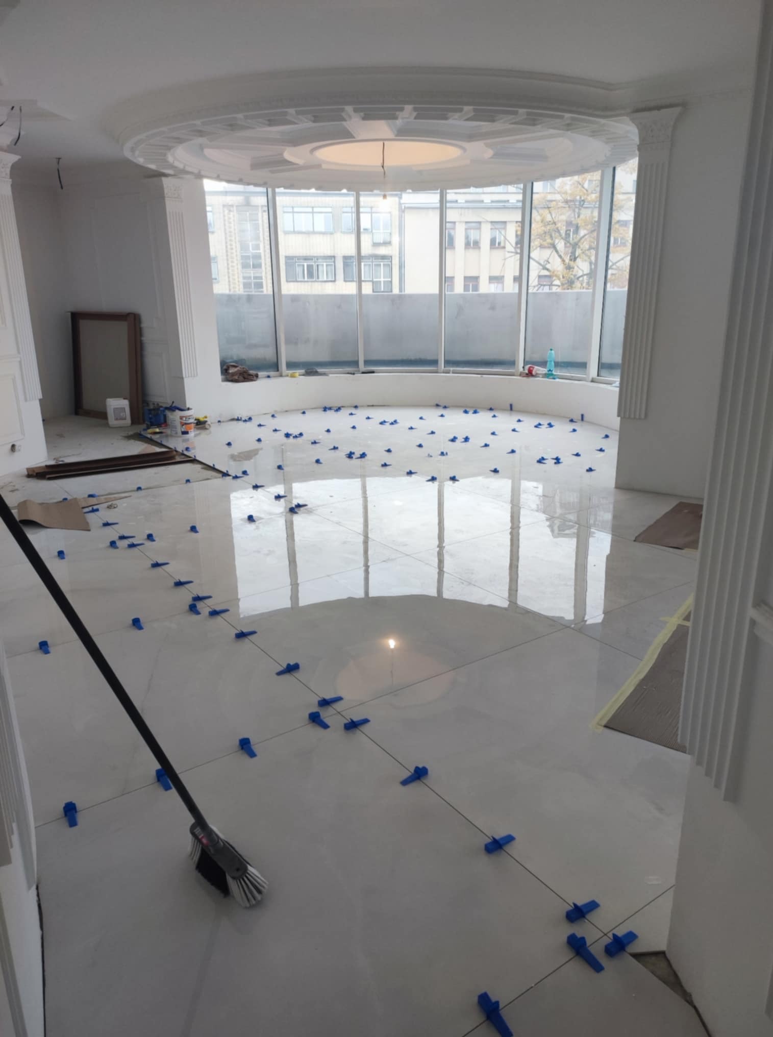 Błyszcząca, jasna podłoga z płytek w trakcie układania, z niebieskimi krzyżykami dystansowymi. Widok na okno i fragment sufitu z dekoracyjnym elementem.