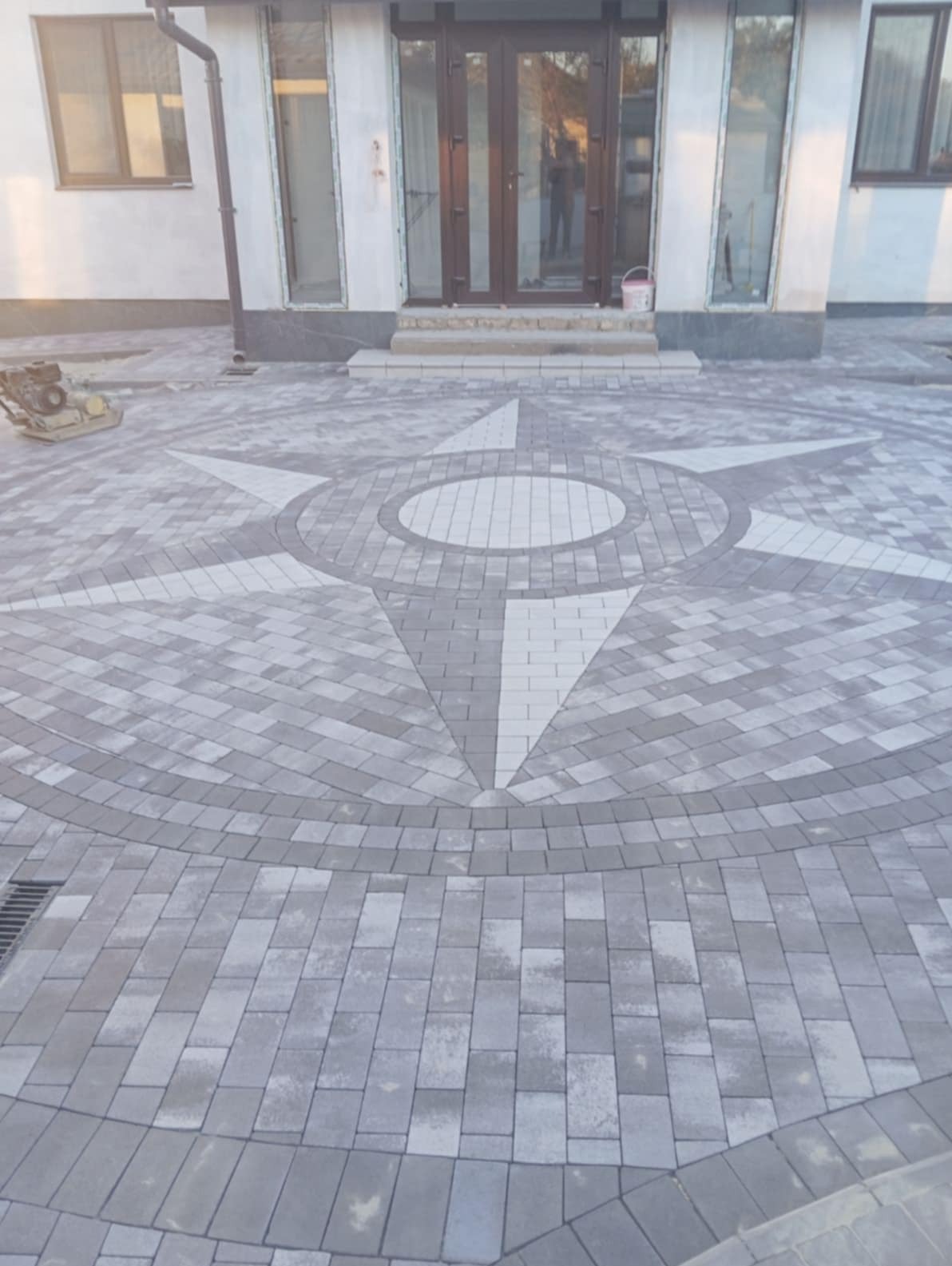 Ułożona kostka brukowa w kształcie rozety przed budynkiem z brązowymi drzwiami. Widoczna zagęszczarka. Szara kostka z białymi akcentami.