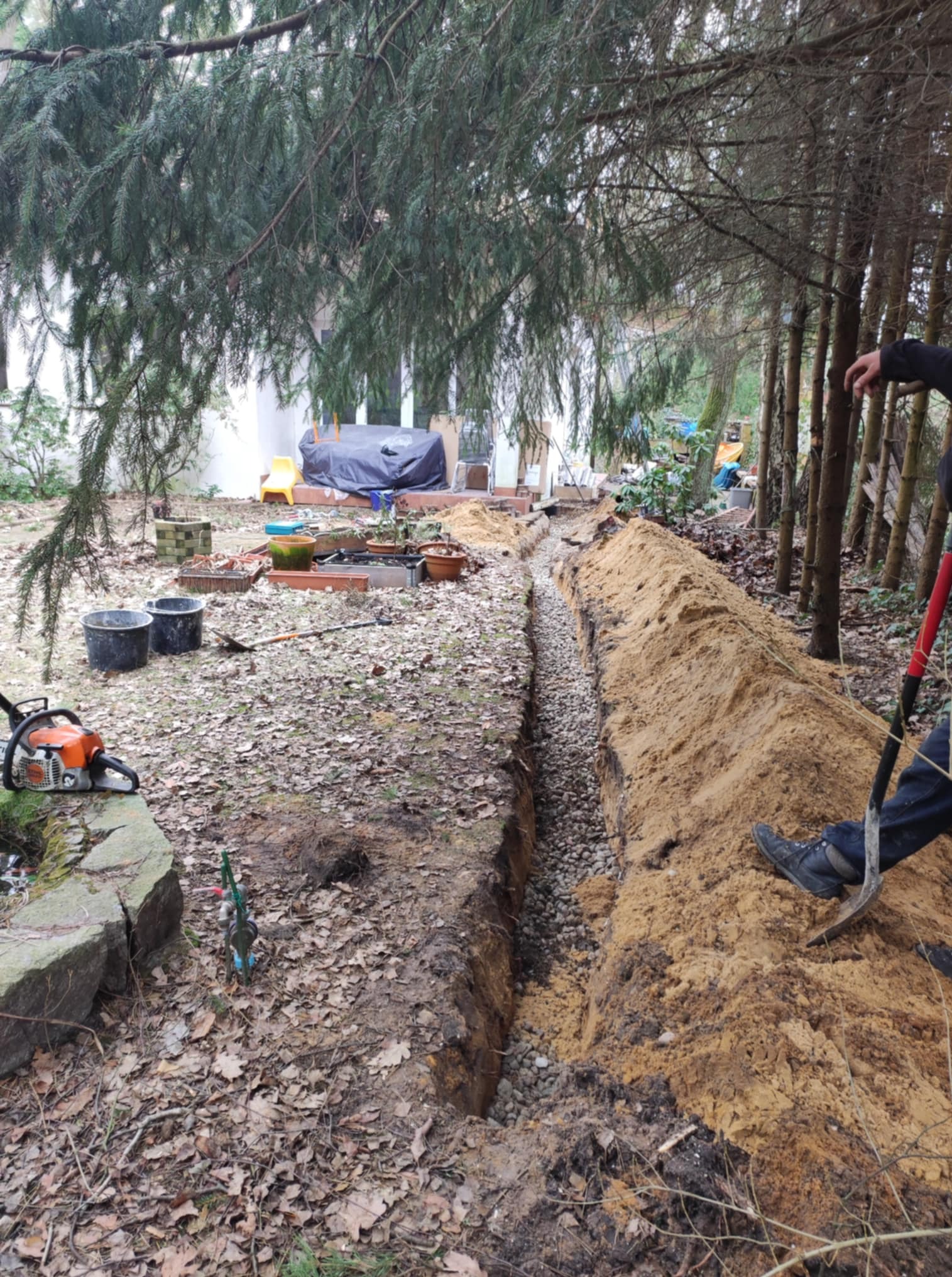 Wykopany rów z drenażem wypełniony żwirem, widoczna ziemia, drzewa i narzędzia. Prace ziemne w ogrodzie, przygotowanie do odprowadzania wody.