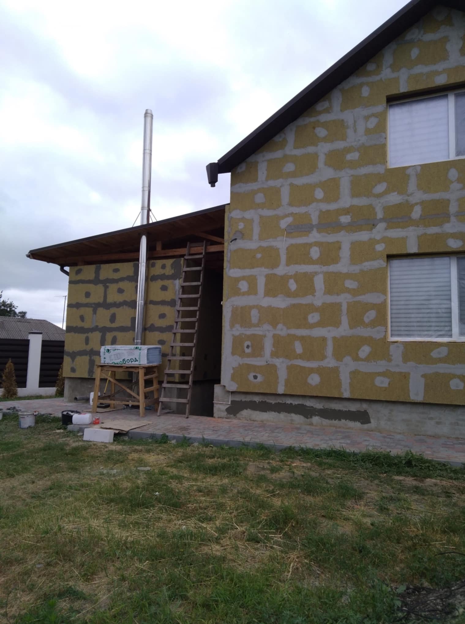 Ocieplanie budynku wełną mineralną, widoczne płyty izolacyjne i rusztowanie. Kominek z blachy wystaje ponad dach. Prace w toku, widok z zewnątrz.