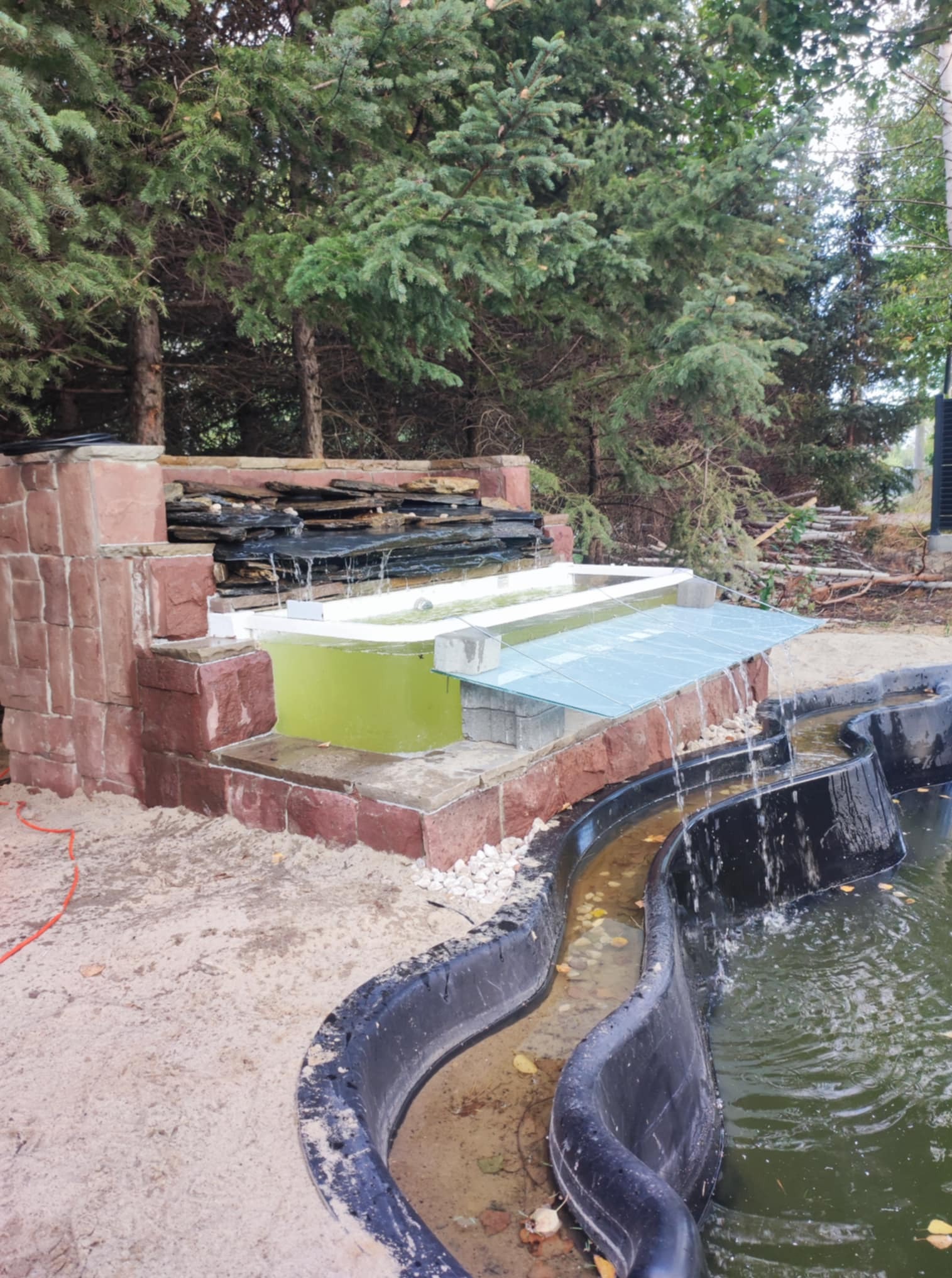 Kaskada wodna z kamienia i zbiornikami wodnymi, otoczona piaskiem i zielenią. Woda spływa po kamieniach do czarnego, falistego stawu. Widoczne drzewa w tle.